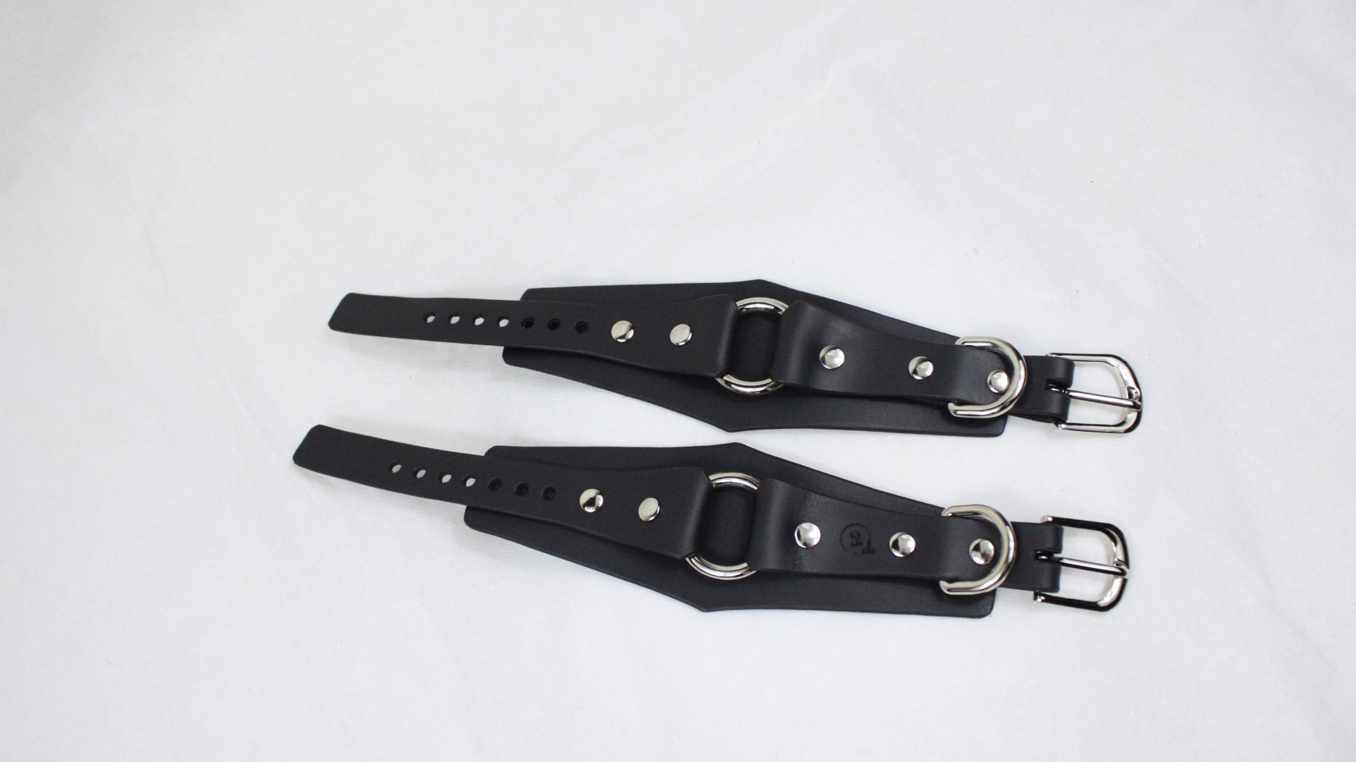 A pair of black leather straps with metal buckles and holes, laid out on a white surface.