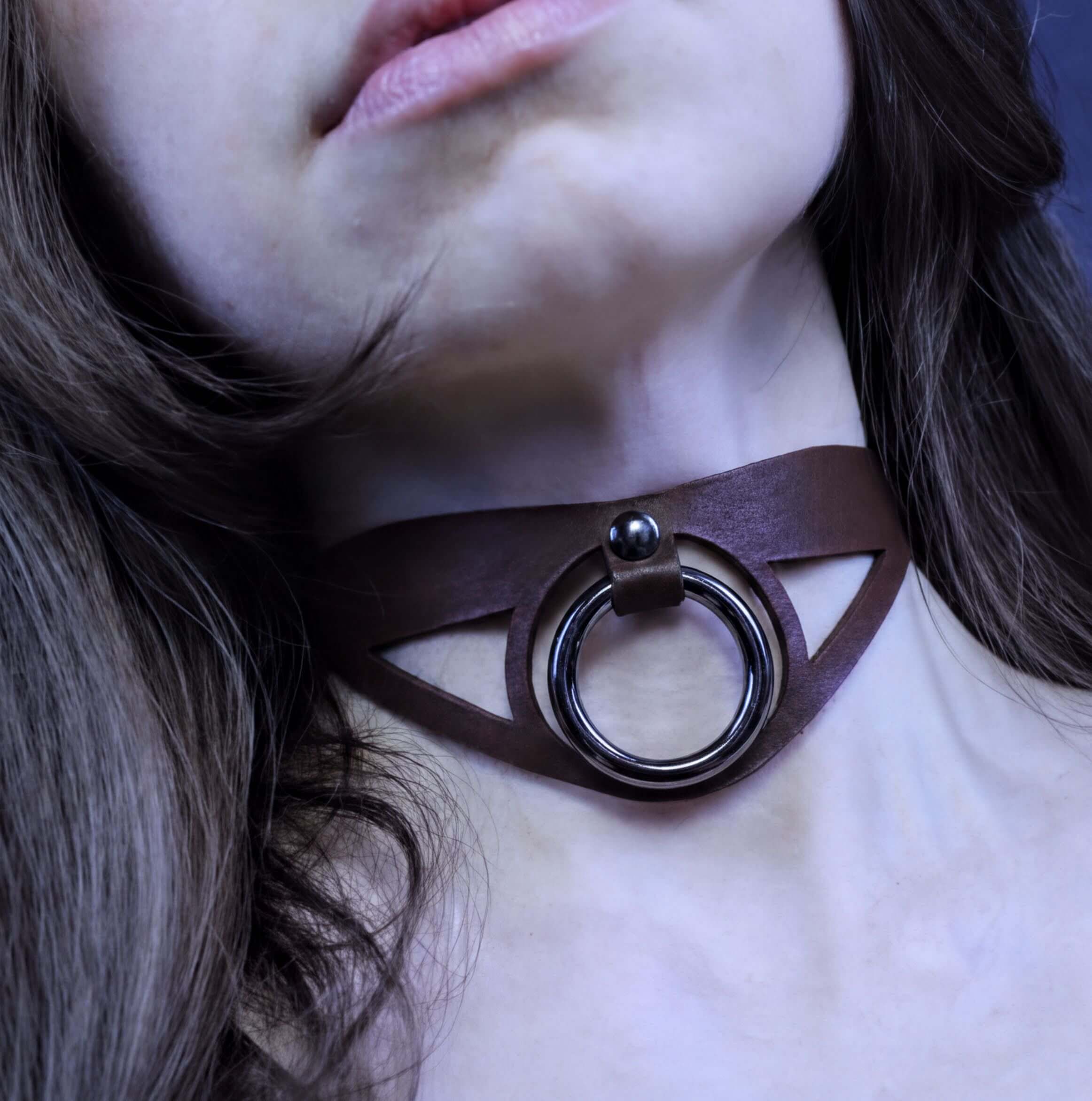 A close-up of a person's neck and chin area, featuring a dark brown choker necklace with a circular metal ring.