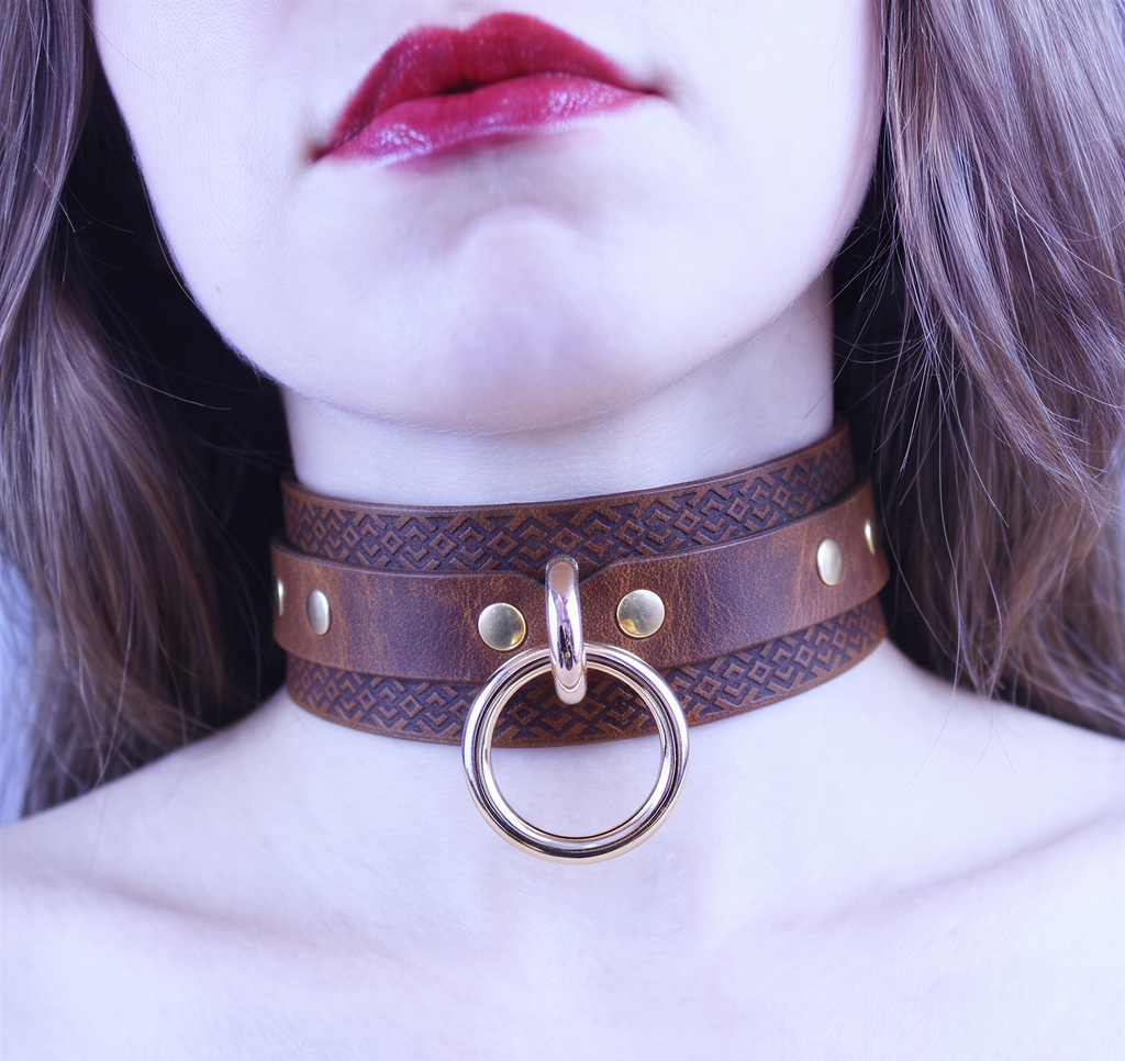 A close-up of a woman's face wearing a brown leather choker necklace with a gold ring and studs.