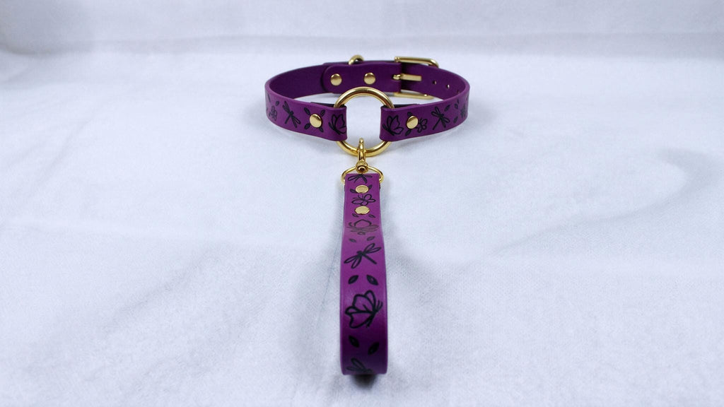 A purple collar with a gold buckle and a black floral pattern on the strap is displayed on a white surface.