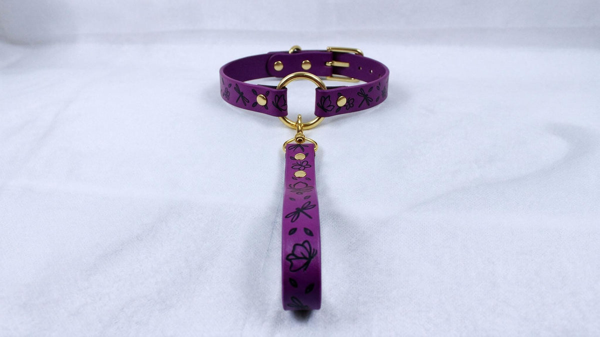A purple collar with a gold buckle and a black floral pattern on the strap is displayed on a white surface.