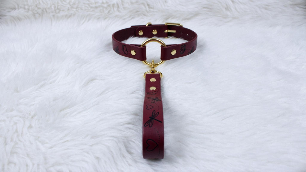 A burgundy leather collar with a gold buckle and handle is displayed on a white furry surface.