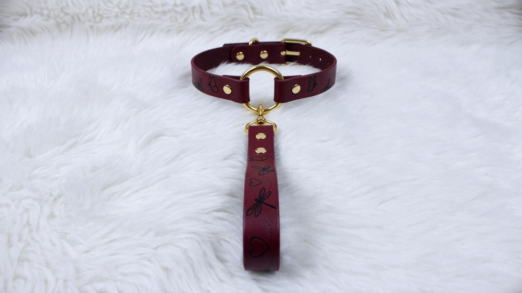 A burgundy leather collar with a gold buckle and handle is displayed on a white furry surface.