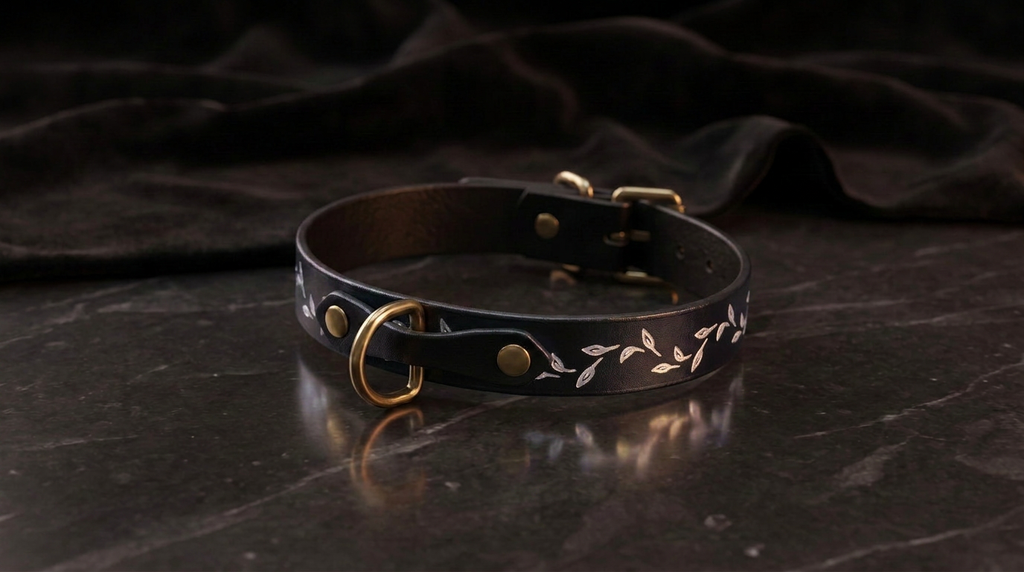 A black leather collar with a gold-colored buckle and leaf design, resting on a dark surface.