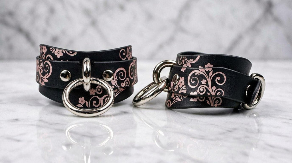 a pair of black leather cinch belts with floral designs and metal rings on the ends, placed on a marble surface.