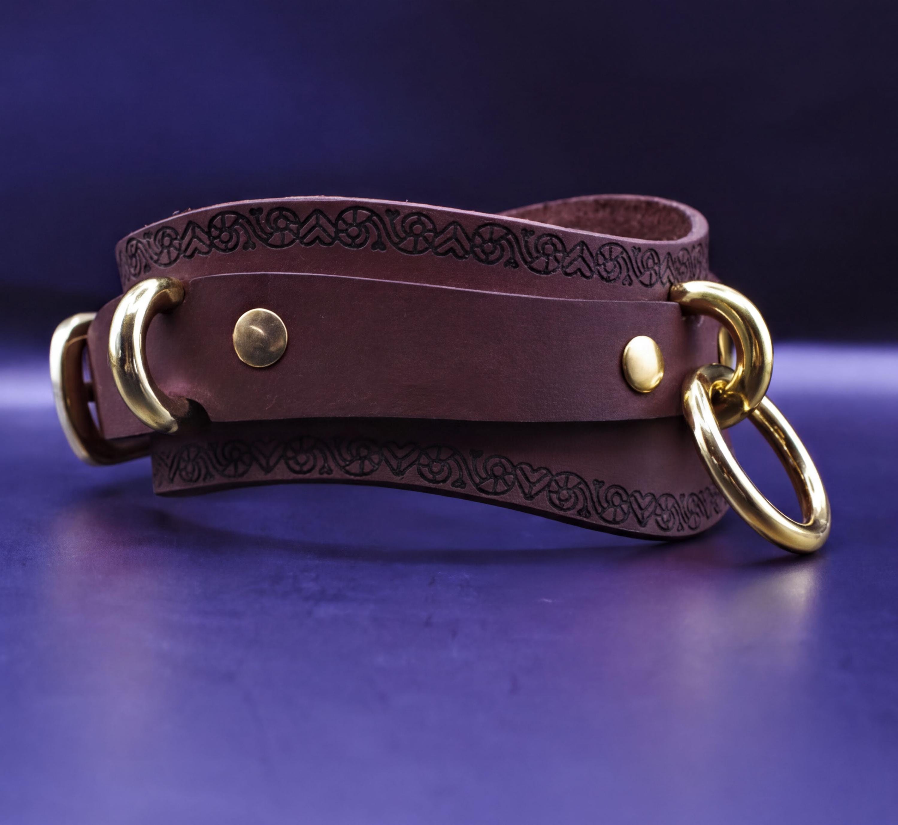A brown leather choker with gold metal rings and a decorative pattern on the strap is displayed against a dark blue background.