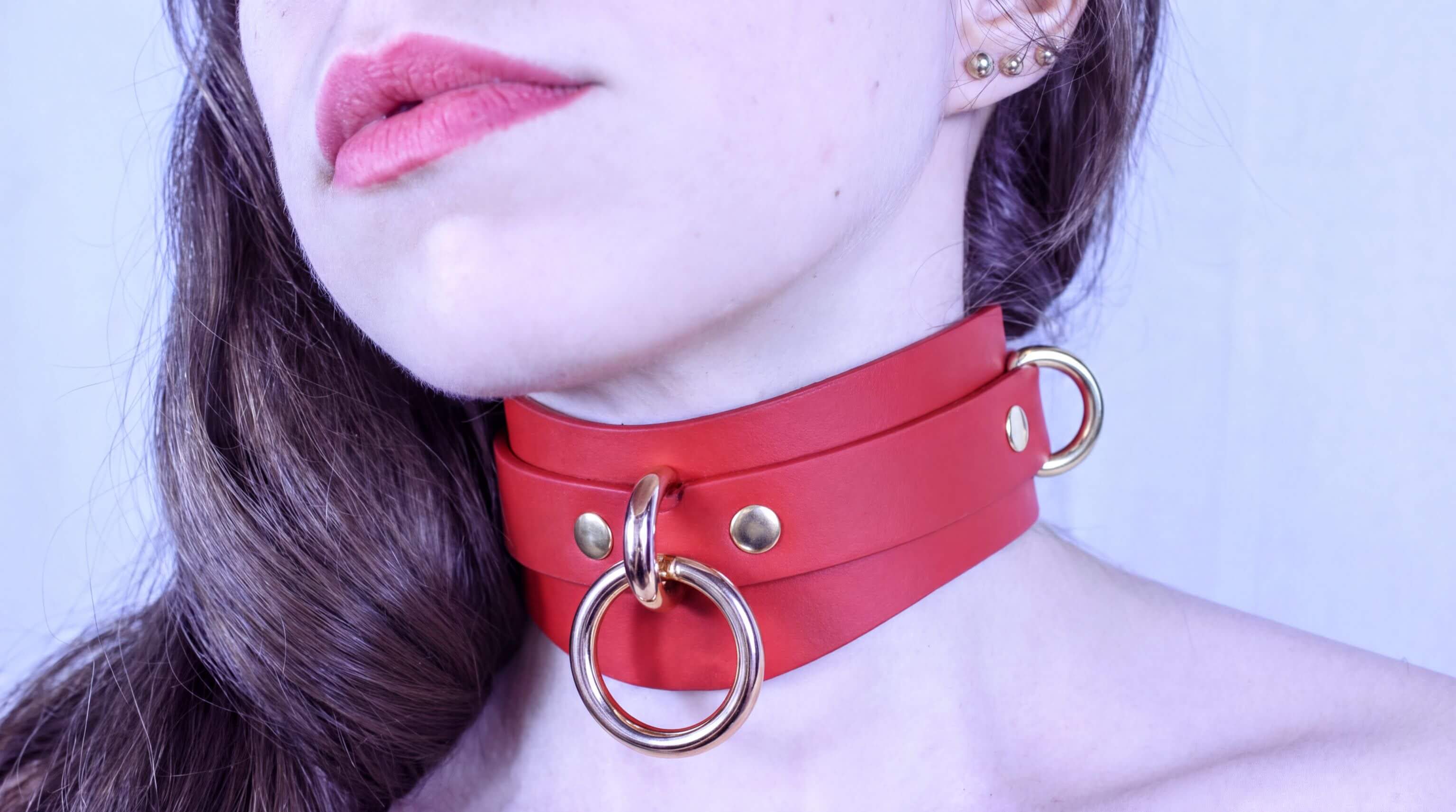 A close-up of a woman's face wearing a red leather choker necklace with gold hardware.