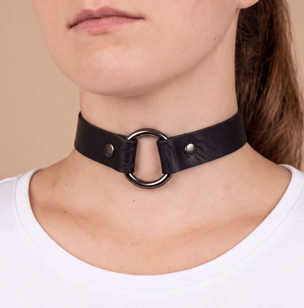 A close-up of a woman's neck wearing a black leather choker necklace with a metal ring.