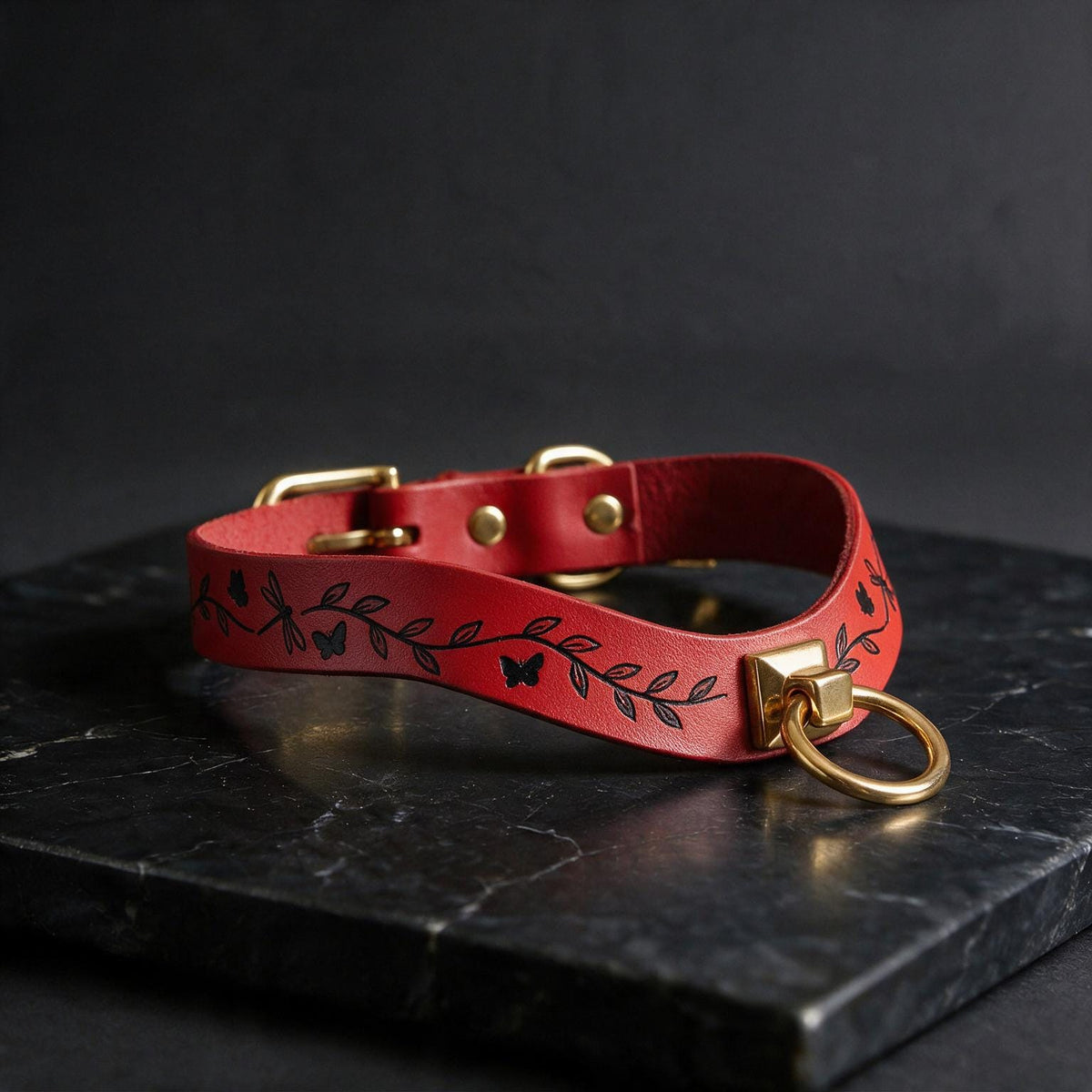 A red leather collar with a leaf and butterfly design, featuring a gold-colored metal ring and buckle, is displayed on a black marble surface.