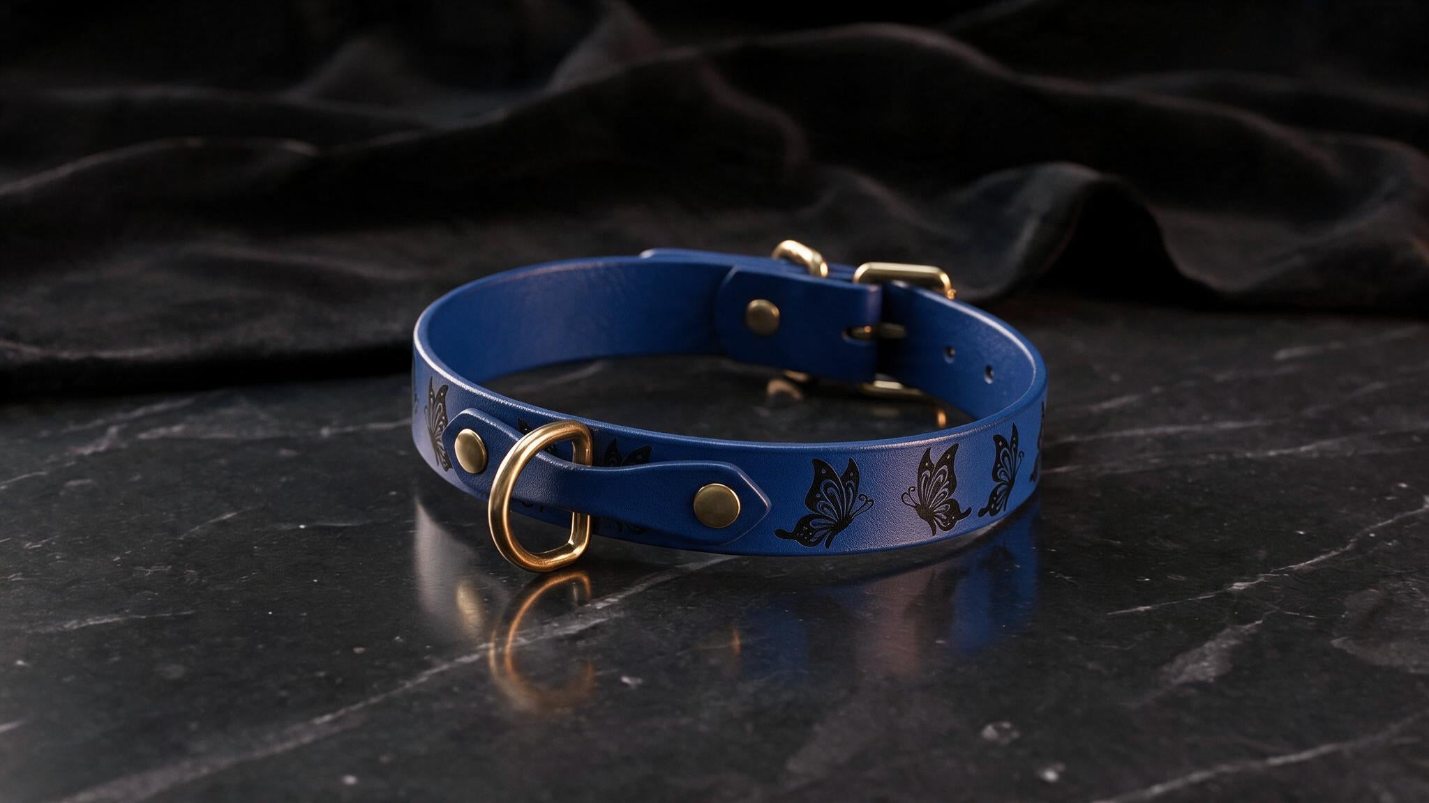 A blue leather dog collar with a gold buckle and butterfly design, placed on a black marble surface.