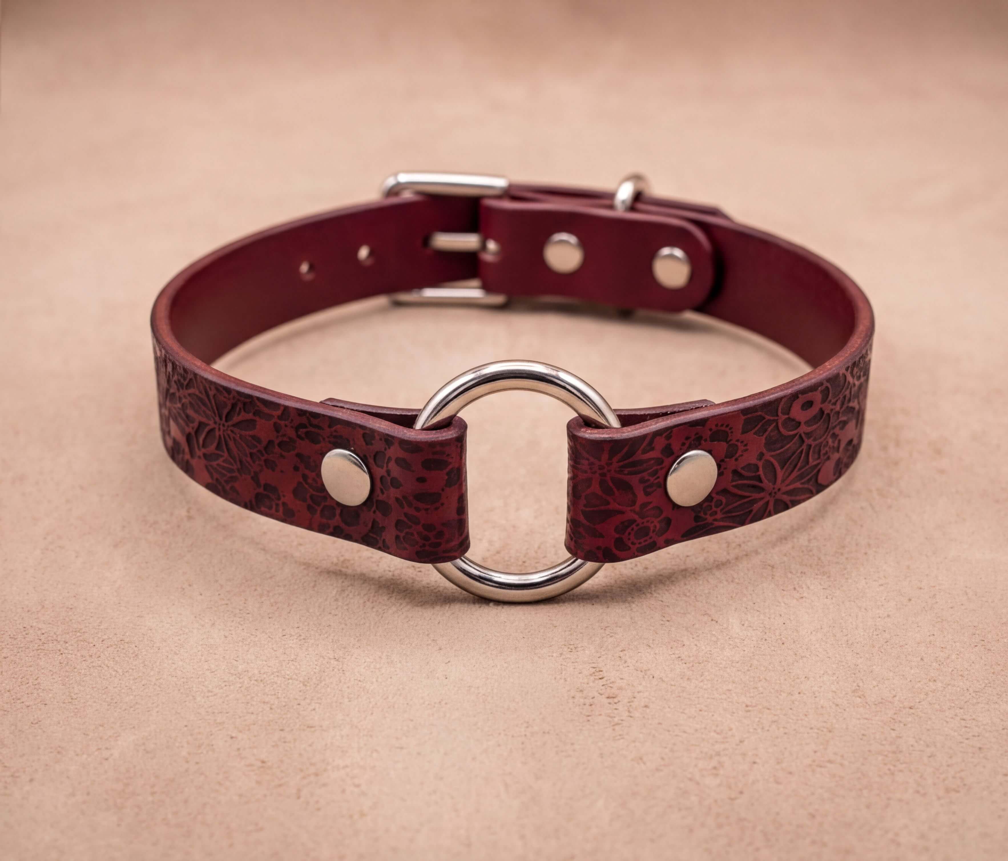 A burgundy leather collar with a metal ring and buckle, displayed on a beige background.