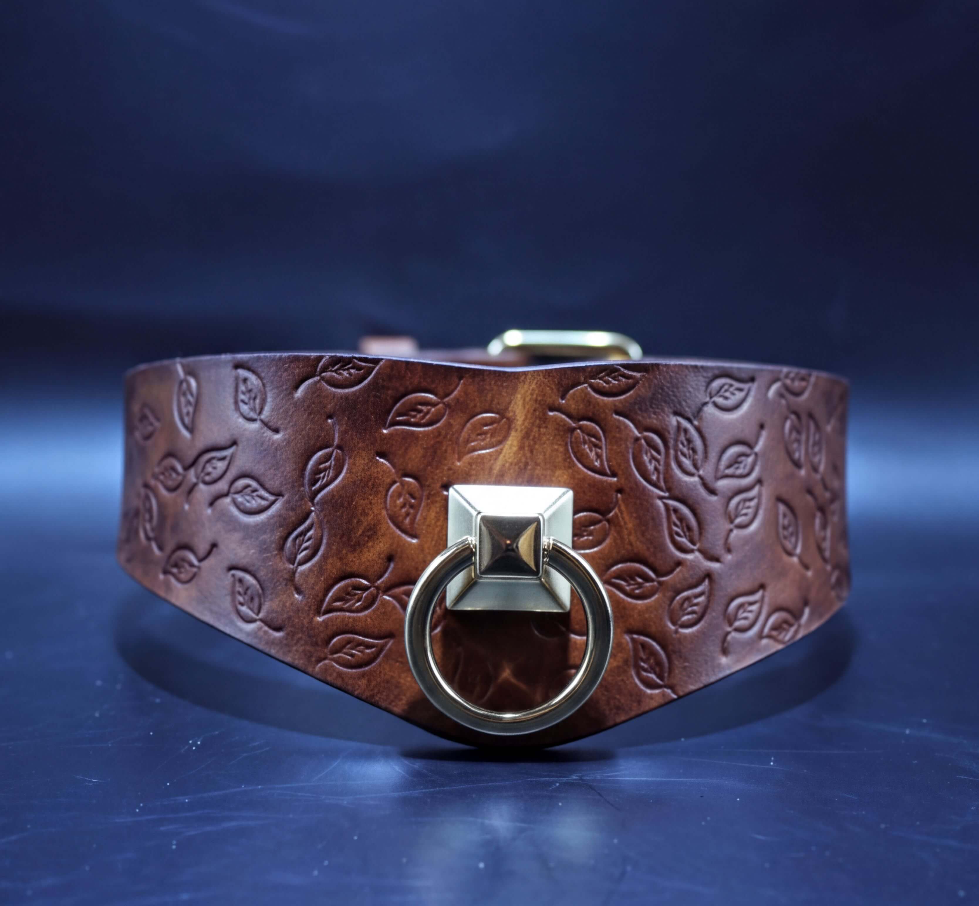 A brown leather collar with a leaf pattern and a gold-colored metal ring with a square-shaped design.