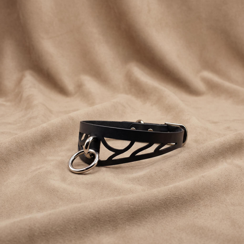 A black leather choker necklace with a metal ring and buckle is displayed on a beige fabric background.