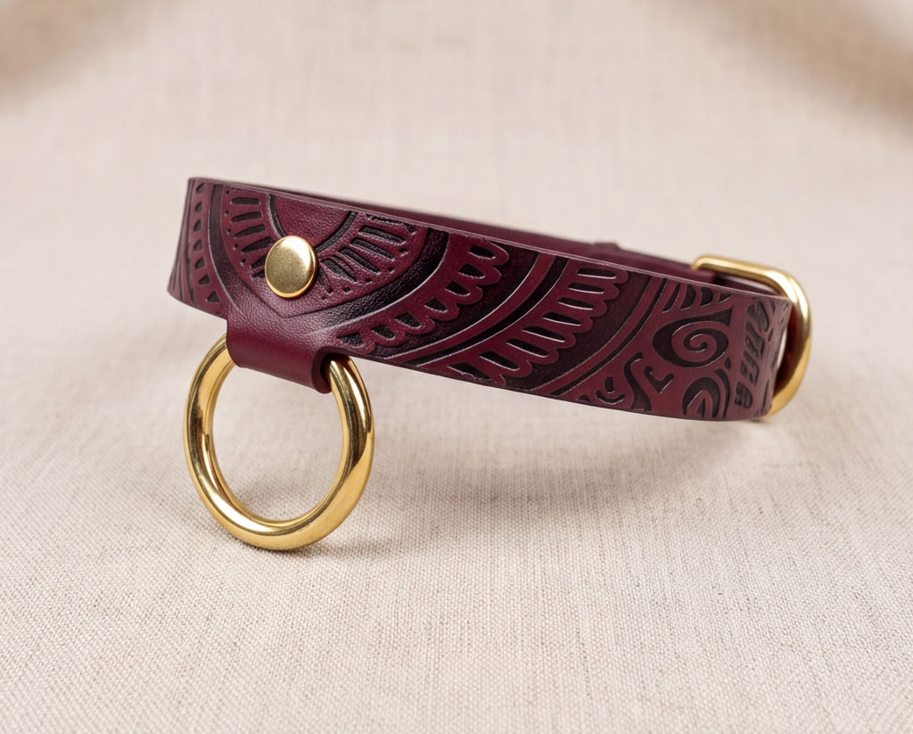A burgundy leather collar with a gold-colored metal ring and buckle, placed on a beige fabric background.