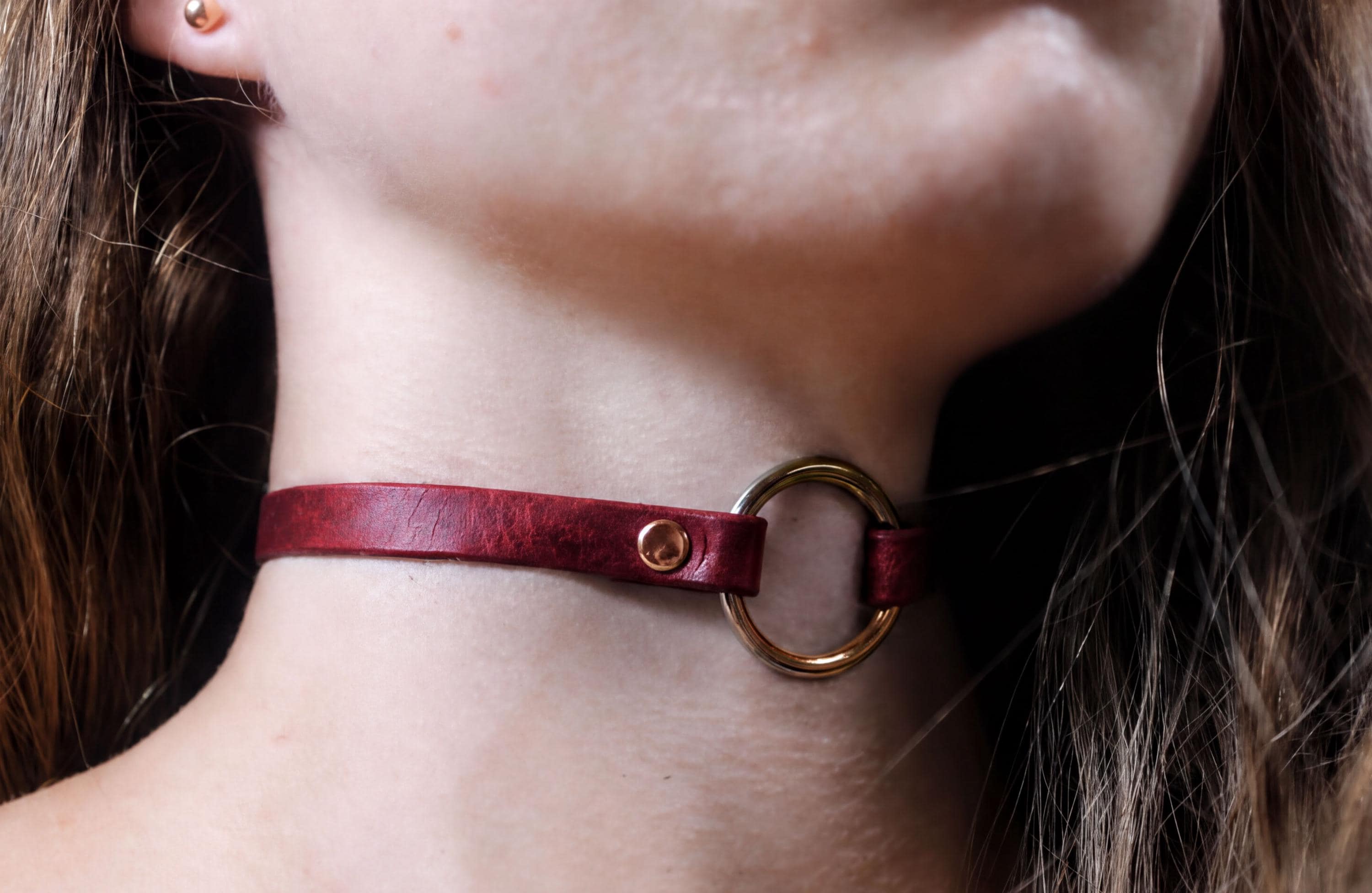 A close-up view of a woman's neck and chin area, featuring a red leather choker necklace with a gold-colored ring.