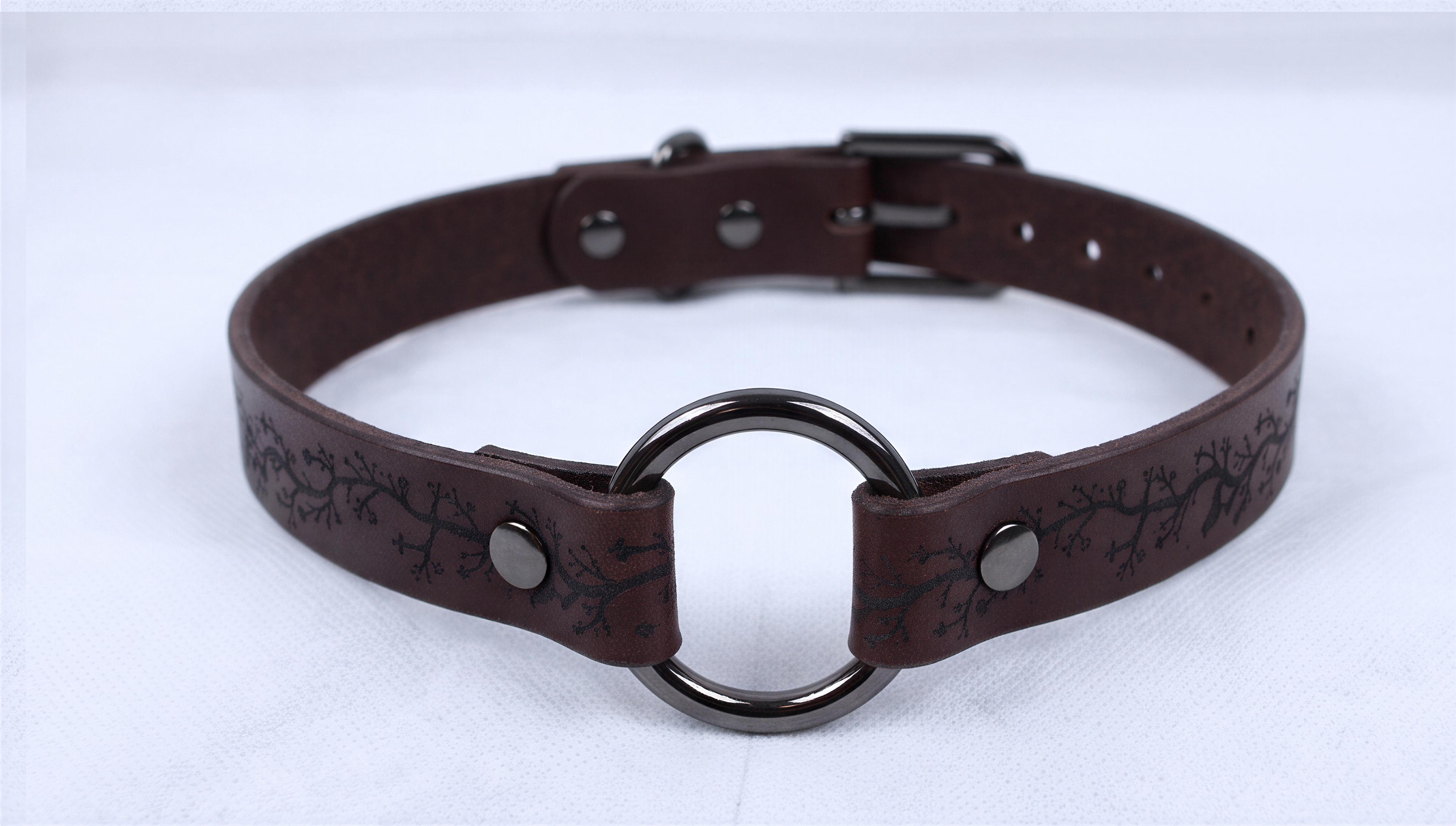 A brown leather choker necklace with a metal ring and a decorative pattern on the leather.