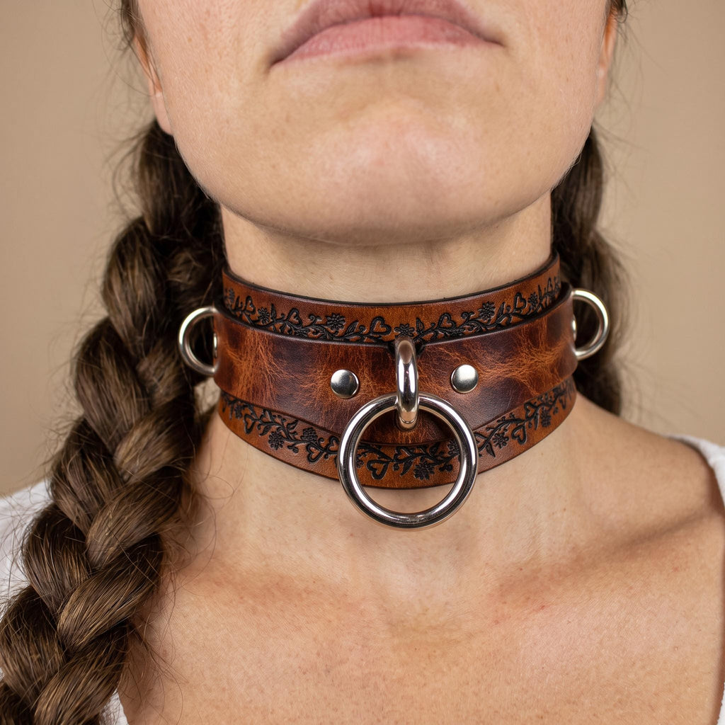 A close-up of a woman's neck and chin wearing a leather choker necklace with metal rings and a circular clasp.
