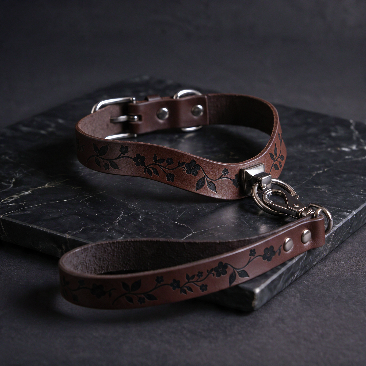 A brown leather collar with a floral pattern and a matching leash attached to it, resting on a dark surface.