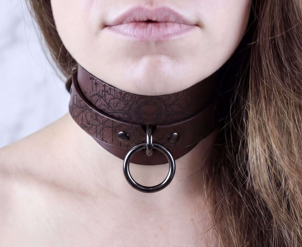 A close-up of a woman's neck and chin area, featuring a dark brown leather choker necklace with a metal ring and buckle.