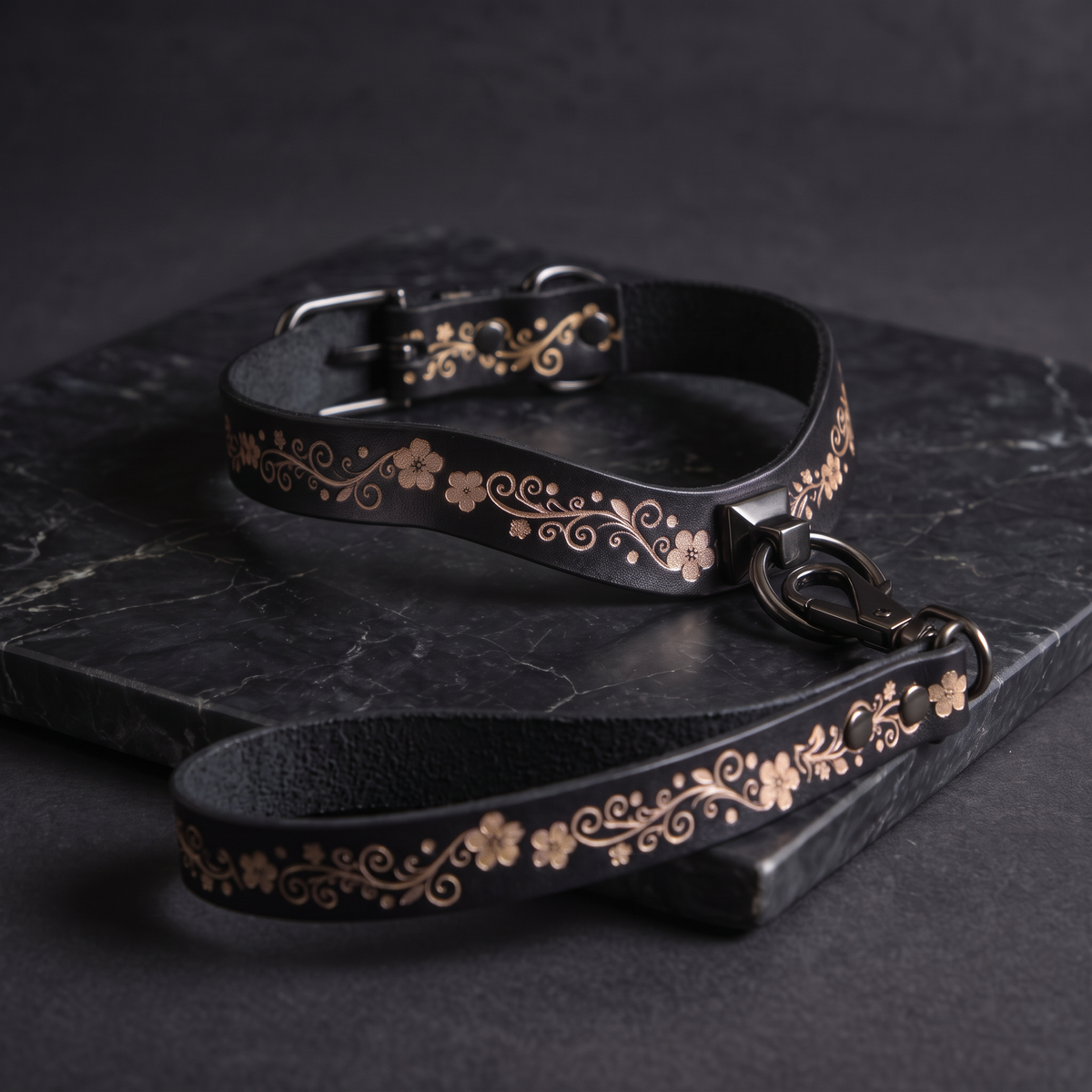 A black leather collar with a floral pattern and a matching leash are displayed on a dark surface.