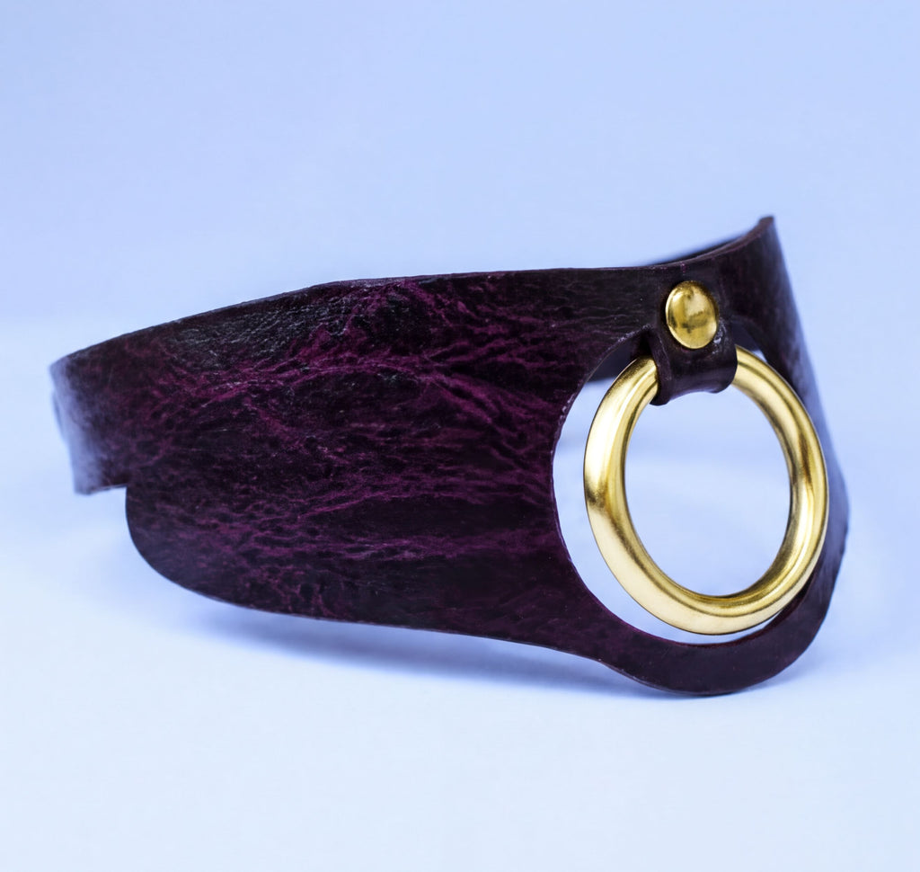 A close-up image of a dark purple leather choker necklace with a gold-colored circular clasp.