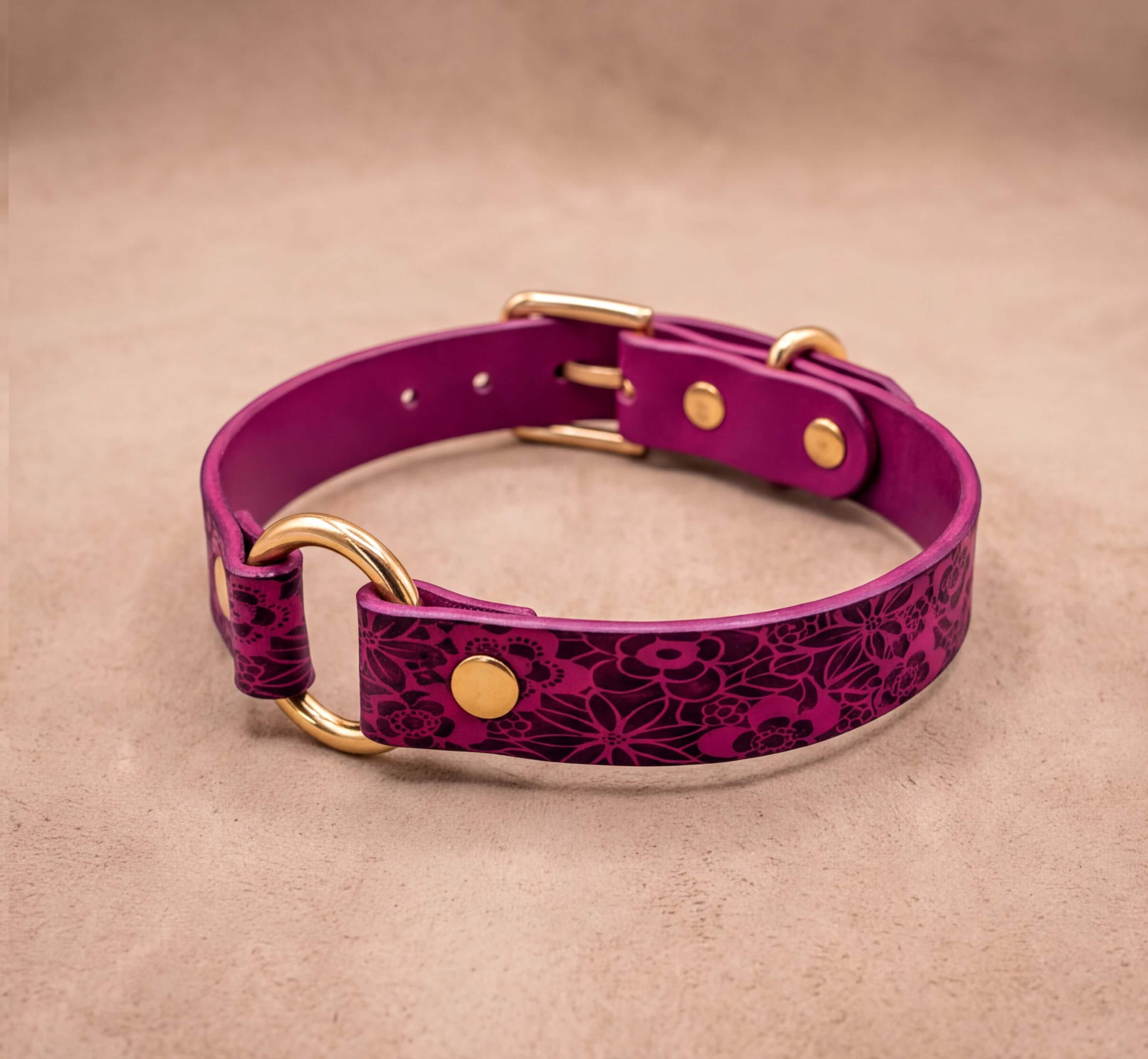 A purple leather collar with a floral pattern and gold hardware, including a buckle and ring, is displayed on a beige background.