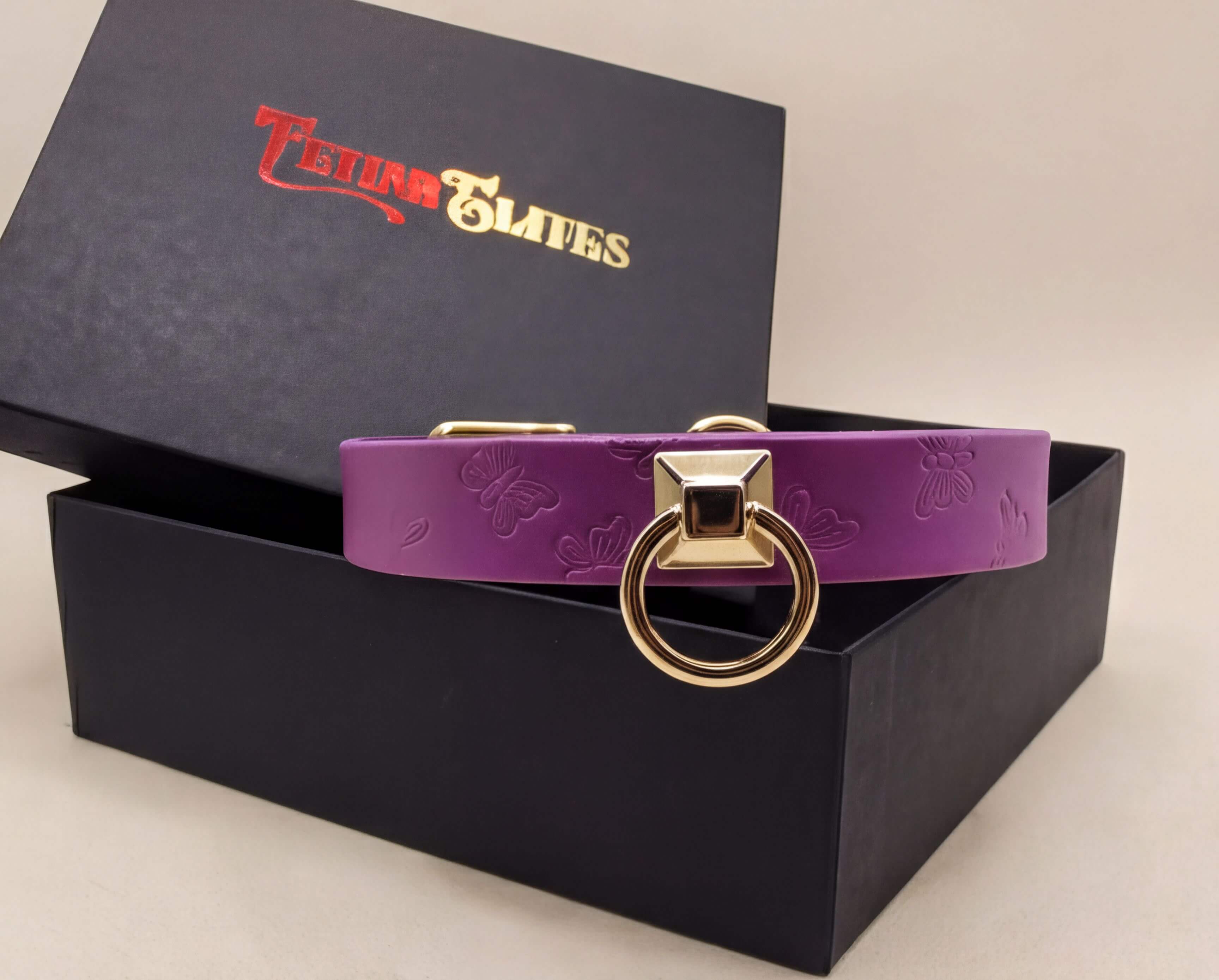 A purple leather collar with a gold-colored metal ring is displayed on top of a black box with a gold-colored logo.