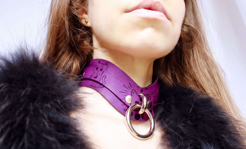 A close-up of a woman wearing a purple choker necklace with a cat design and a large metal ring.