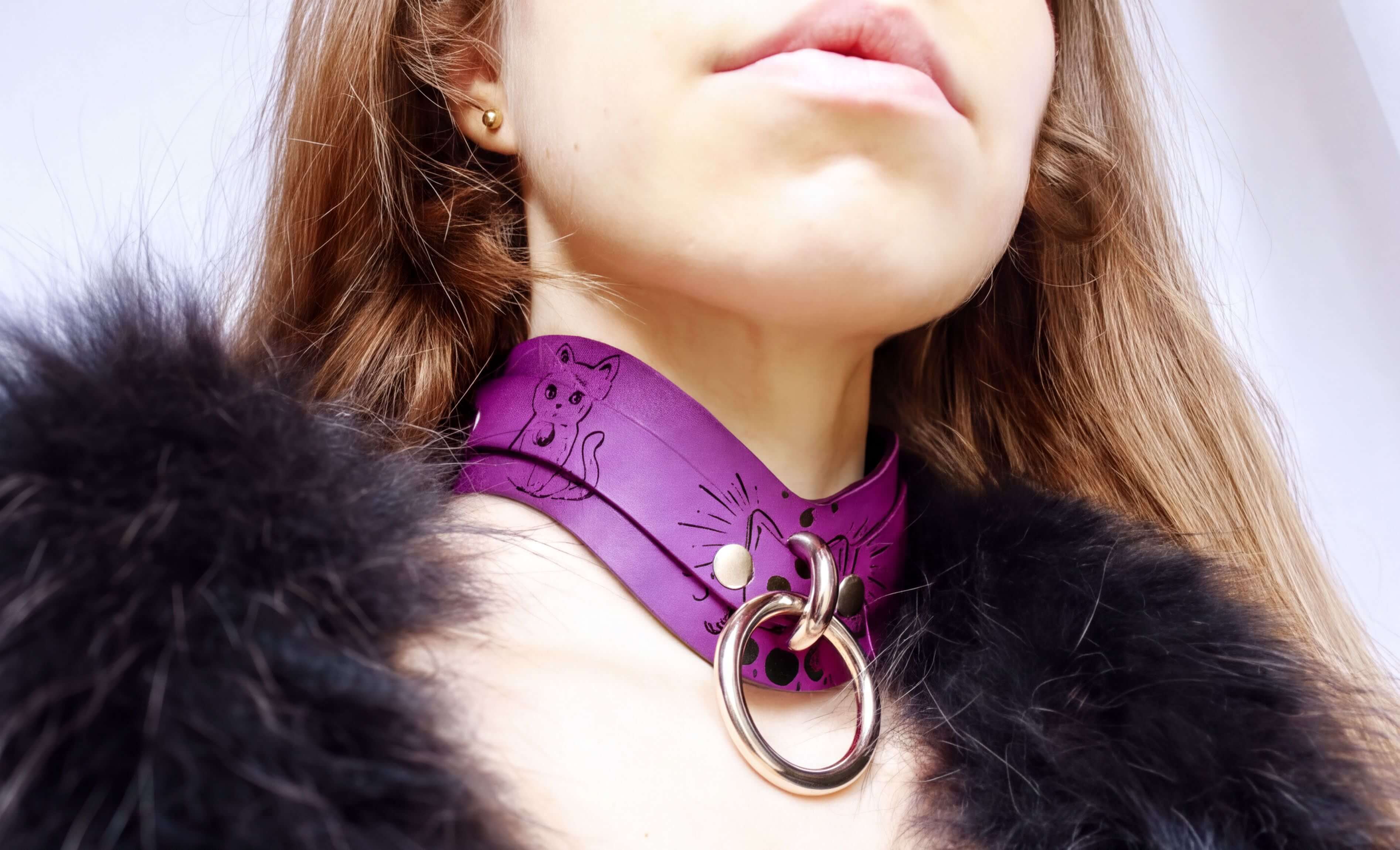 A close-up of a woman wearing a purple choker necklace with a cat design and a large metal ring.