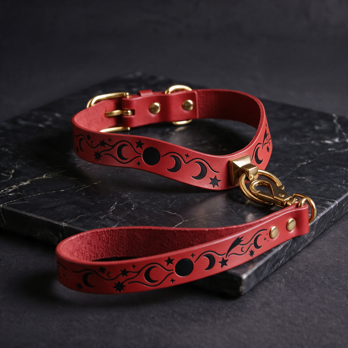 A red leather dog collar with a black star and crescent design, and a matching red leash with a gold buckle, both resting on a black marble surface.