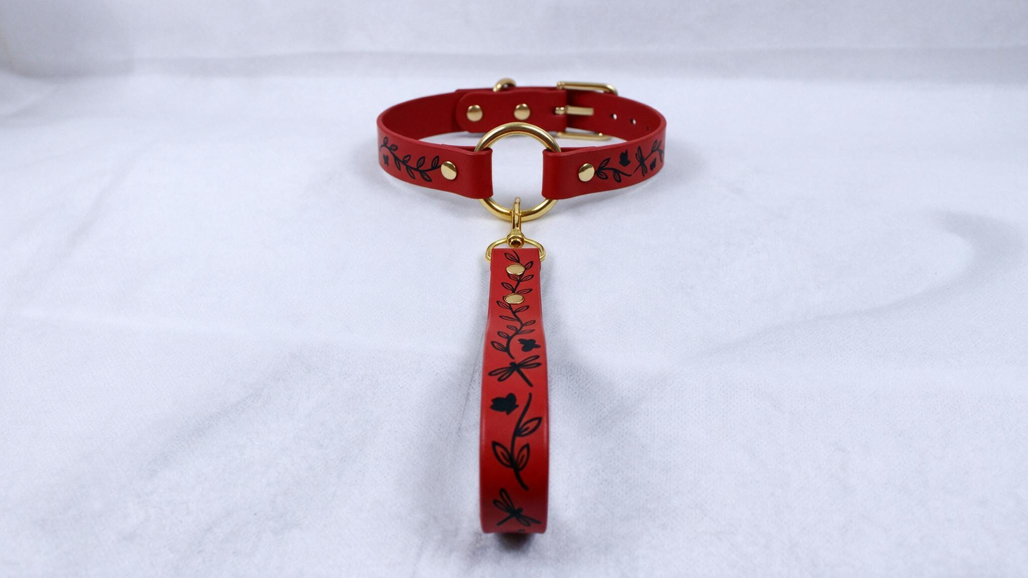 A red leather collar with a gold-colored buckle and a black floral pattern on the strap is displayed against a white background.