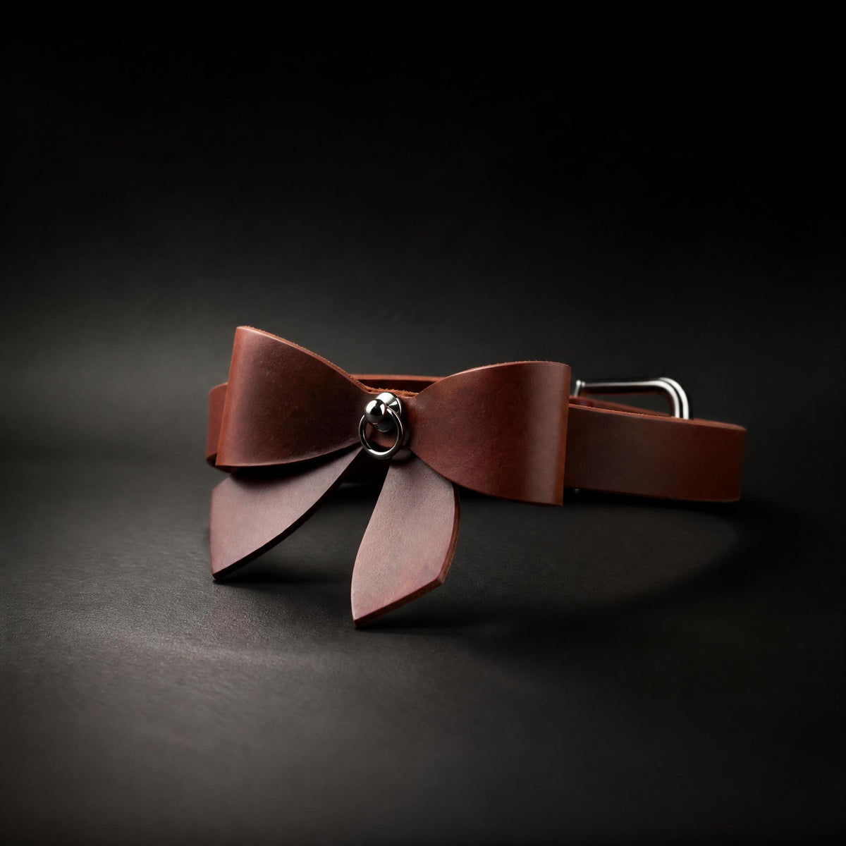 A brown leather bow tie with a silver buckle is displayed against a dark background.