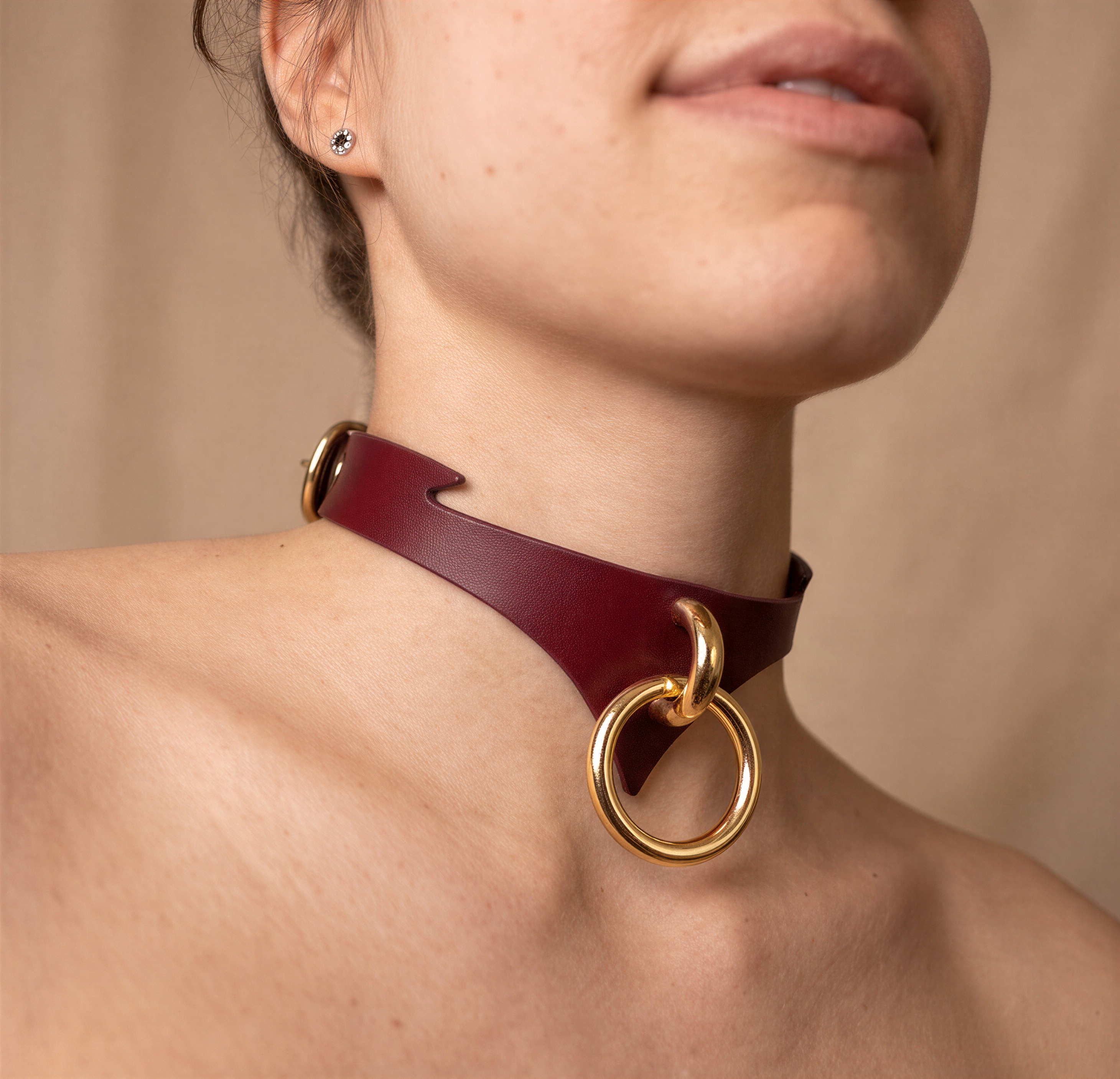 A close-up of a woman's neck and chest area wearing a burgundy leather choker necklace with gold-colored rings.