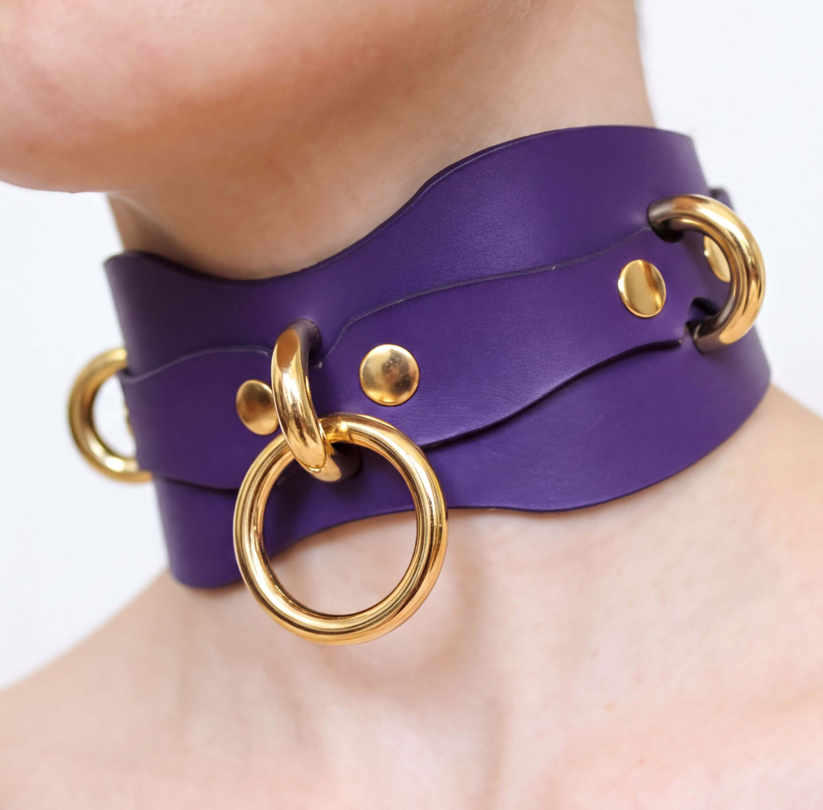 A close-up view of a person's neck wearing a purple leather choker necklace with gold-colored metal rings.