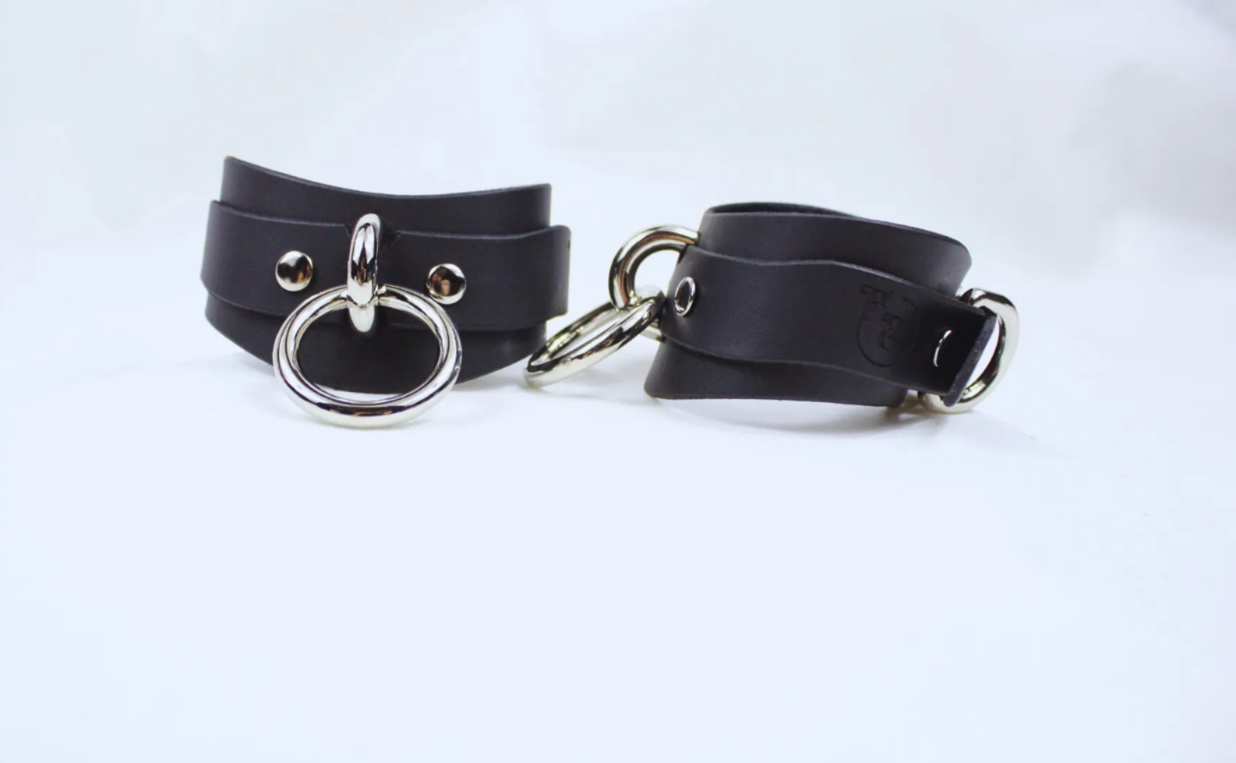 A pair of black leather cuffs with metal rings and buckles, placed on a white background.