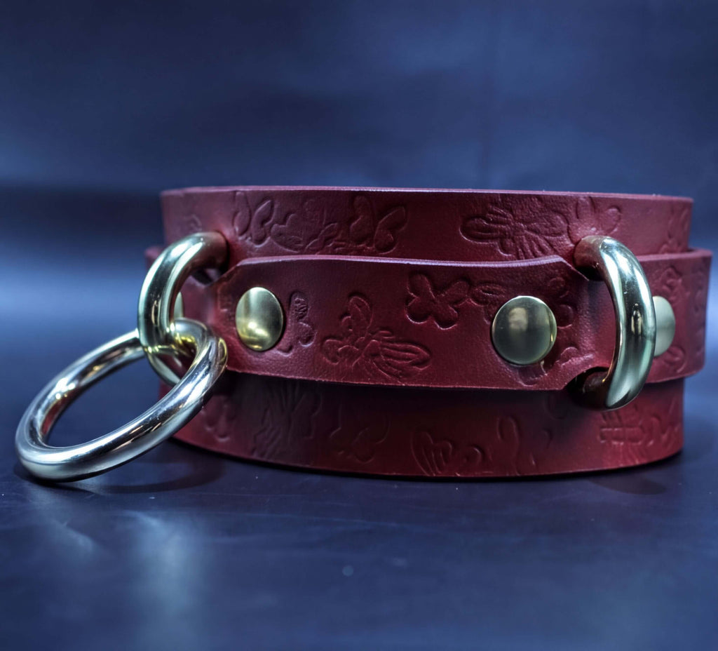 A red leather strap with embossed floral designs and two gold-colored metal rings on the ends, resting on a dark blue background.
