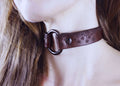 A close-up of a person's neck and chin wearing a brown leather choker necklace with a spider web design.