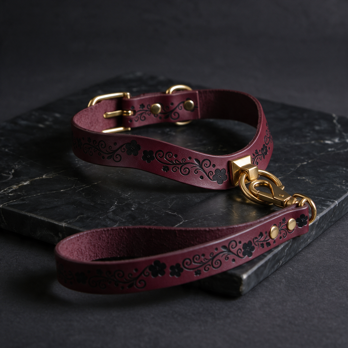 A burgundy leather dog collar with a gold-colored buckle and a matching leash is displayed on a black marble surface.