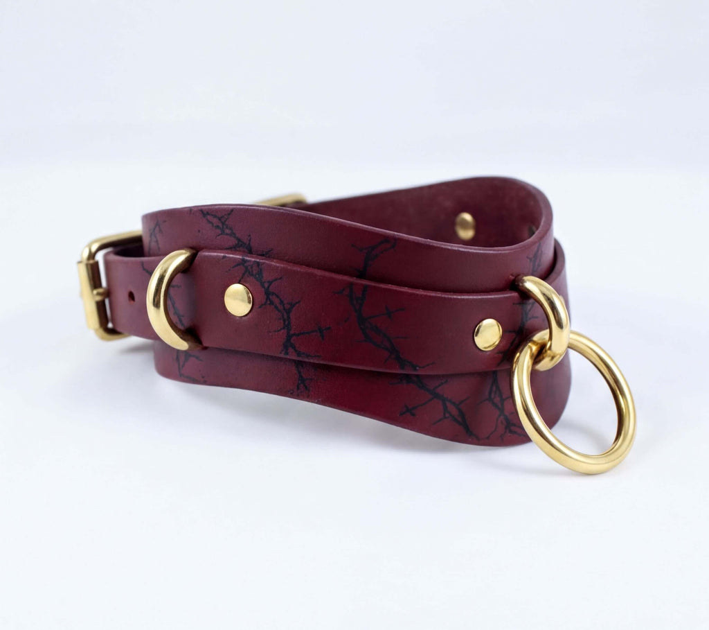 A burgundy leather choker necklace with a gold-colored metal ring and two gold-colored metal rings on the ends.