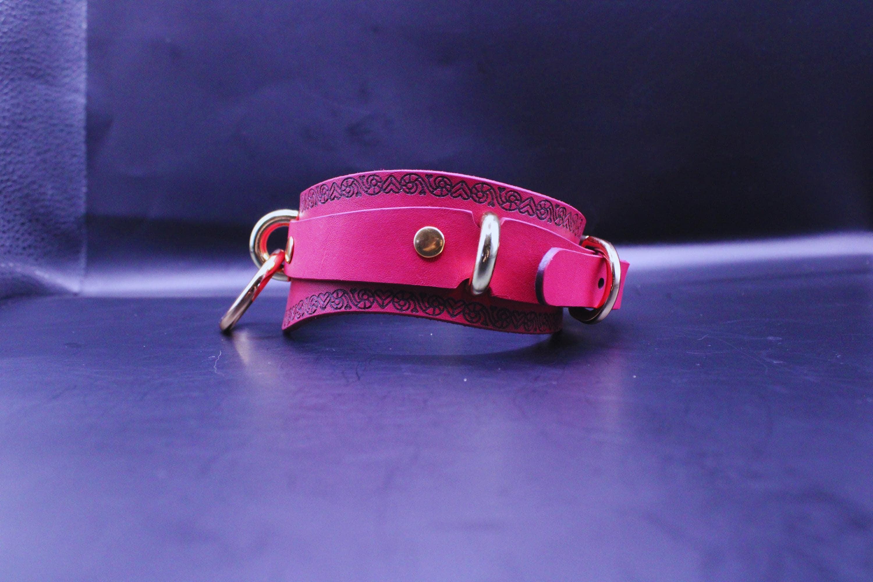 A pink leather collar with metal rings and buckles is displayed on a dark surface.