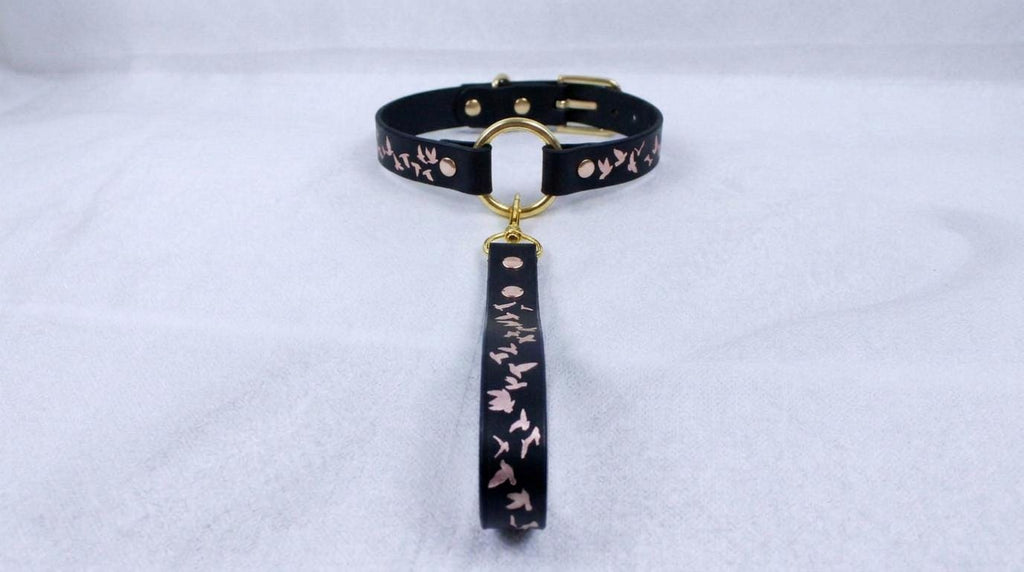 A black leather collar with a gold-colored metal buckle and a dangling tag, displayed on a white surface.