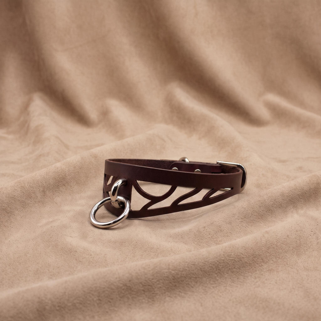 A brown leather choker necklace with a metal ring and buckle is displayed on a beige fabric background.