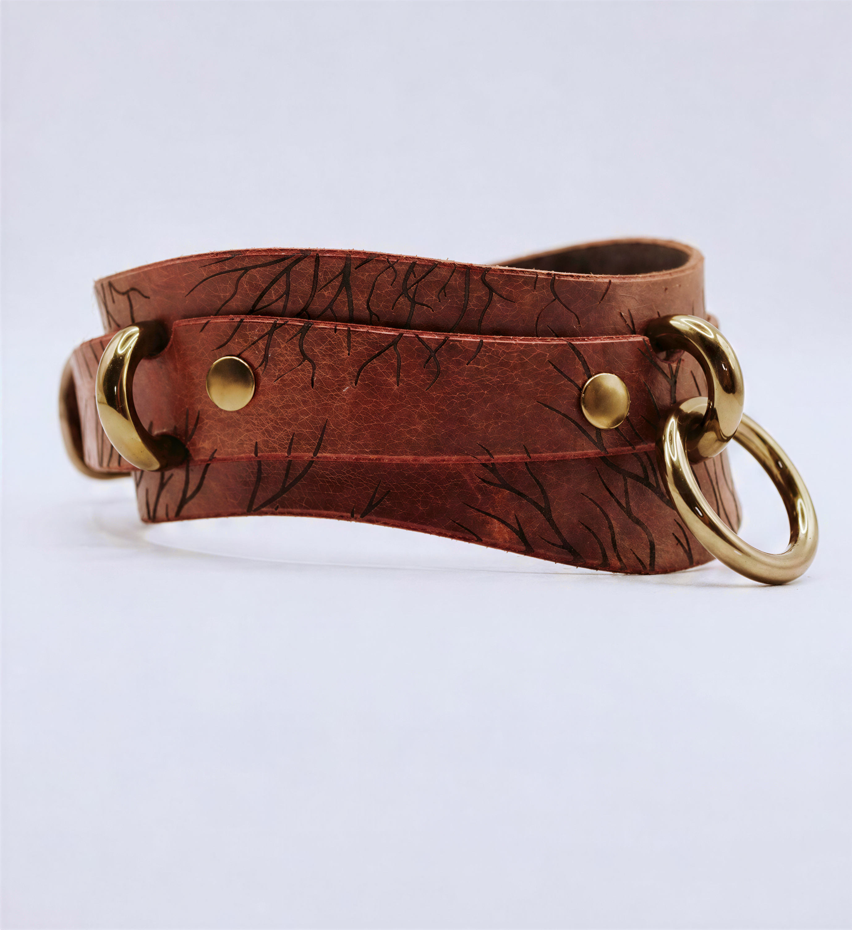 A brown leather strap with a metal ring and two brass buckles, featuring a distinctive tree-like pattern.