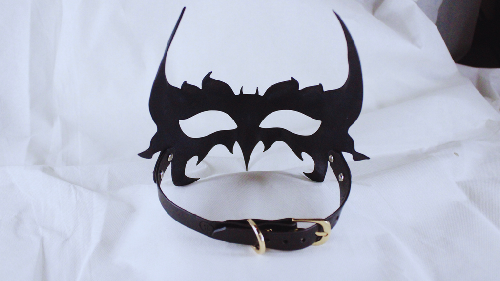 A black mask with a decorative design and a black leather collar is displayed against a white background.