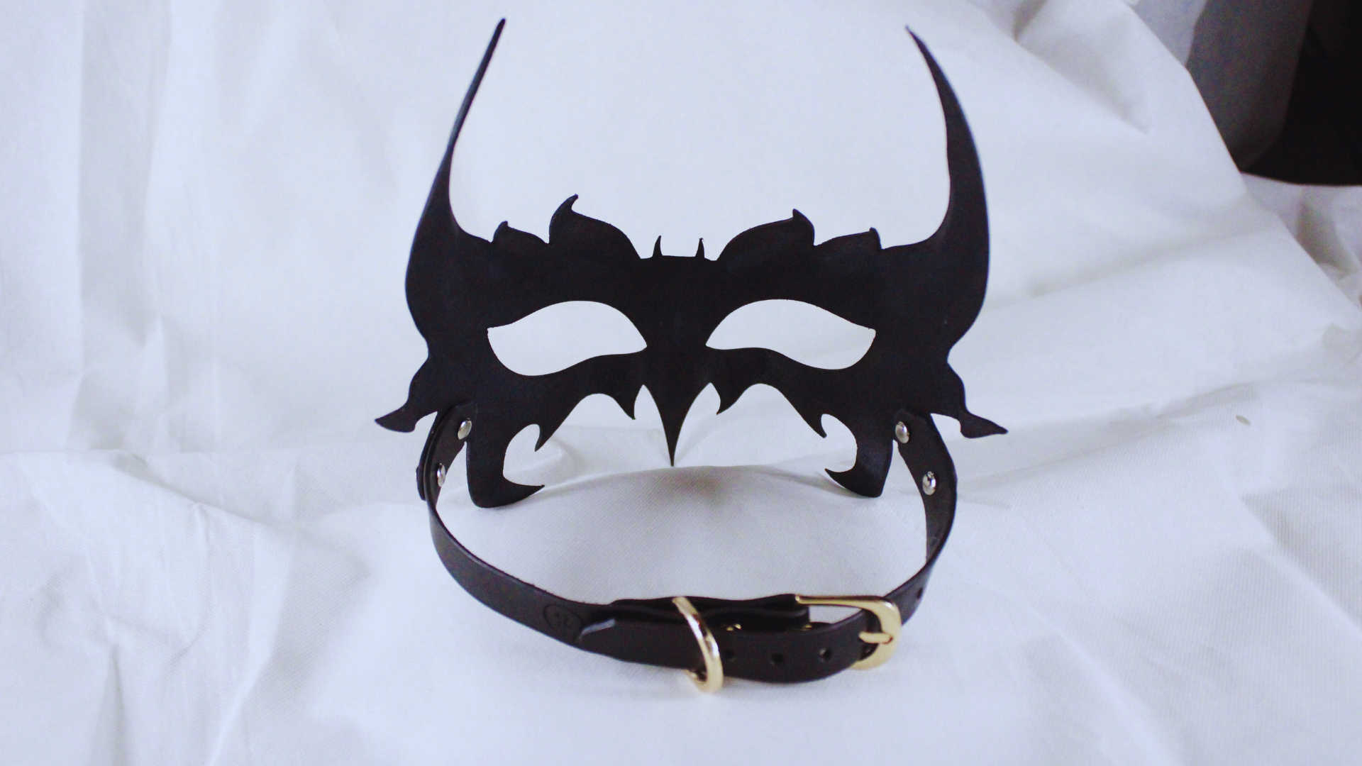 A black mask with a decorative design and a black leather collar is displayed against a white background.