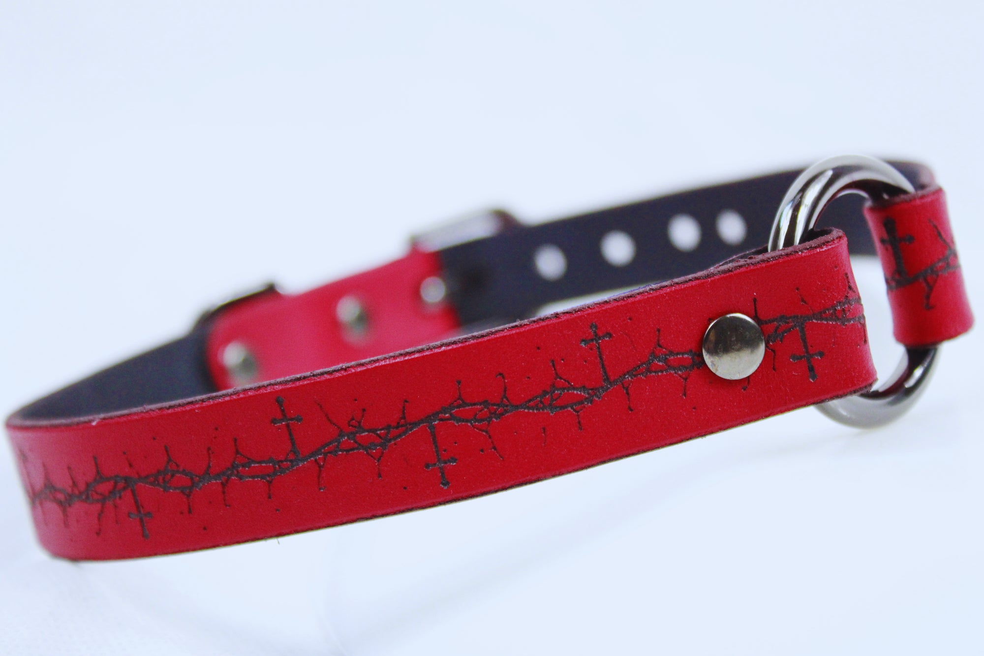 A red leather dog collar with a metal ring and a decorative pattern of crosses on the strap.
