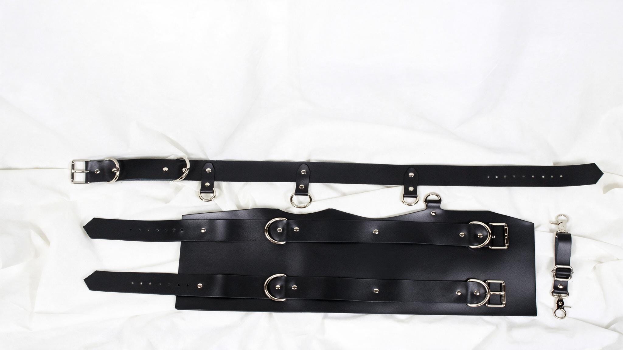 a collection of black leather straps and buckles, including a belt and a harness, laid out on a white background.