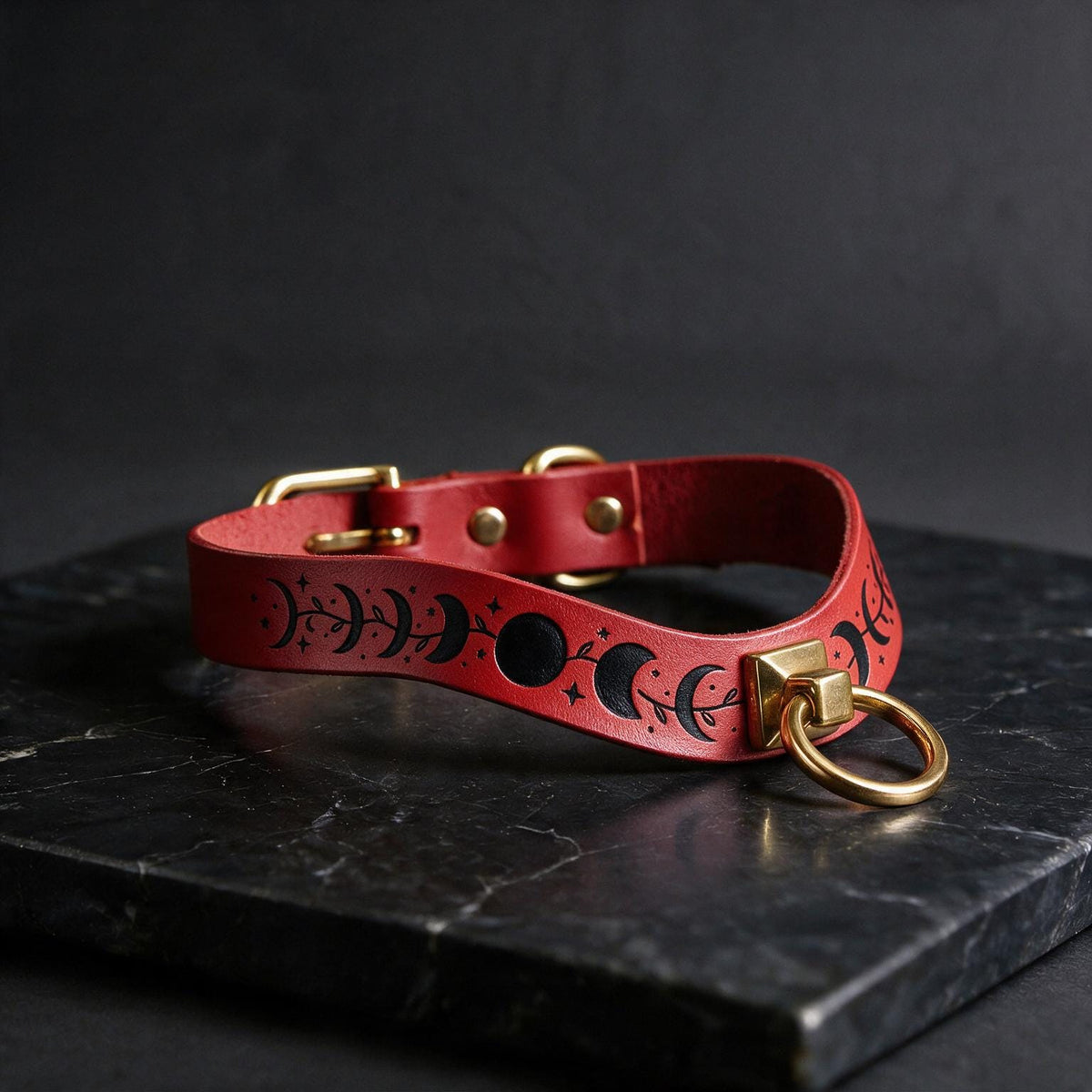 A red leather collar with a moon and star design, featuring a gold-colored metal ring and buckle, is displayed on a black marble surface.