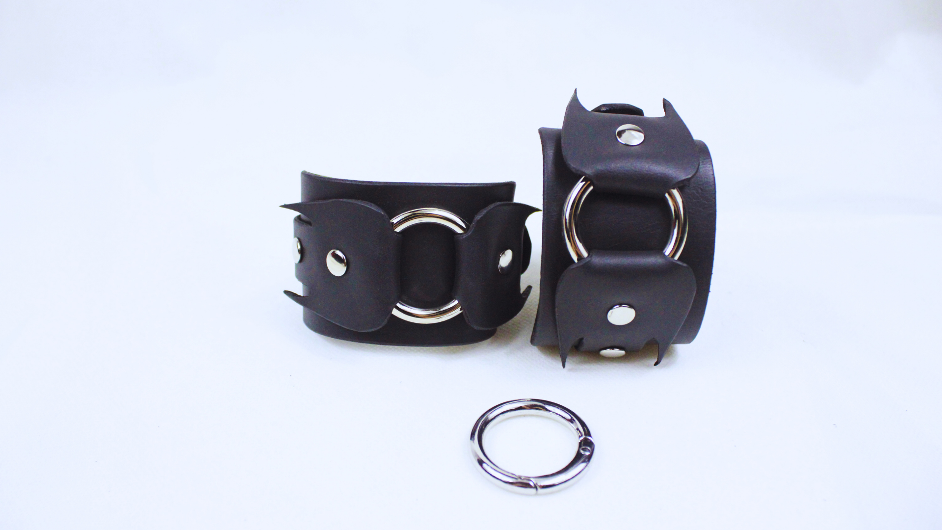 a pair of black leather cuffs with metal rings and a silver ring clasp, placed on a white background.