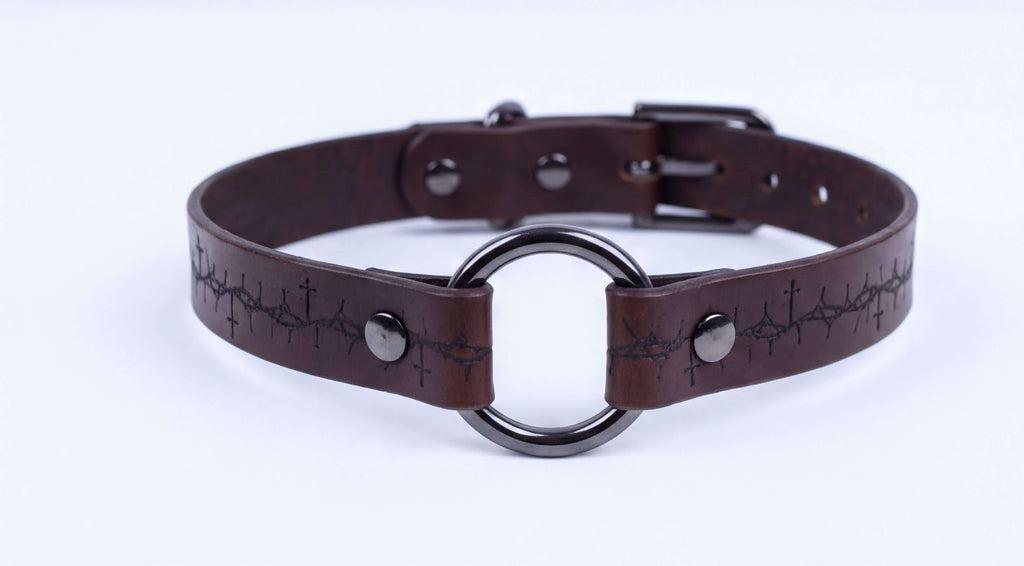 A brown leather choker collar with a metal ring and a cross pattern on the strap.